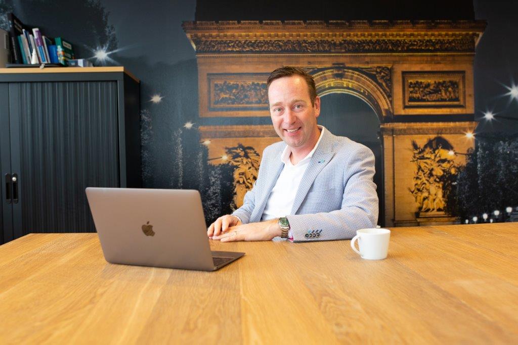 Man in lichtblauw colbert aan houten tafel met laptop en koffie, glimlacht. Foto van Arc de Triomphe hangt aan de muur.