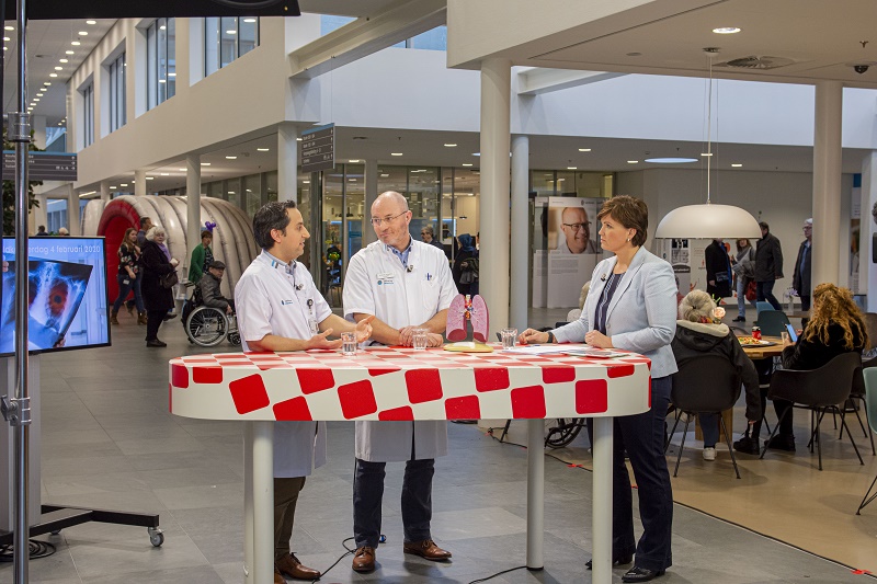 Drie volwassenen bij een rood-wit geblokte tafel bespreken immunotherapie en longkanker; mensen lopen of zitten op de achtergrond.