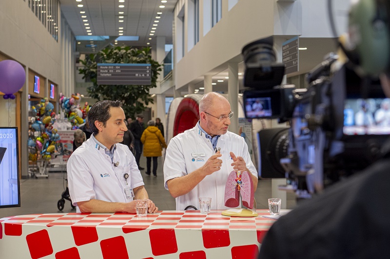 Twee mannen in witte uniformen bespreken longkanker aan een rood-wit geruite tafel. Mensen, ballonnen en info over roken en therapie.