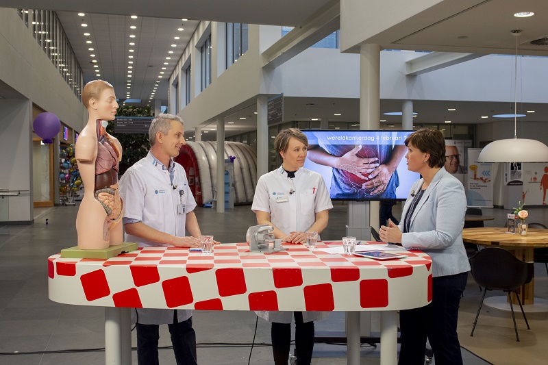 Drie mensen praten aan een tafel met rood-wit kleed in modern gebouw; medisch model en scherm tonen darmkankerbehandeling.