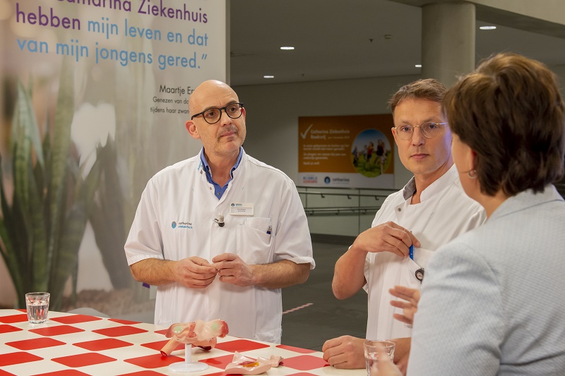 Twee zorgverleners in witte jassen praten met een vrouw bij een tafel; banner met 'Expert Gynaecologische kanker' op achtergrond.