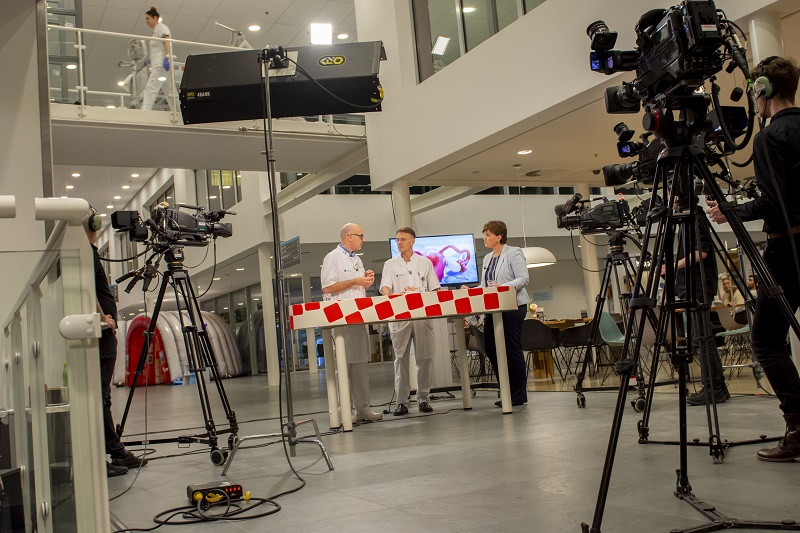 Drie mensen geven een interview over een preventie-app aan een rood-wit gestreepte tafel, met camera's en studiolampen erbij.