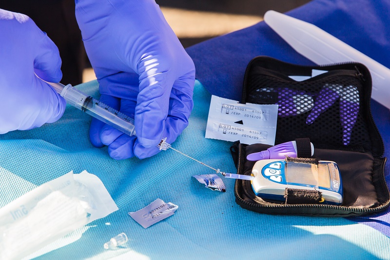 Iemand met paarse handschoenen bereidt een injectiespuit voor bij een glucosemeter en medische benodigdheden.