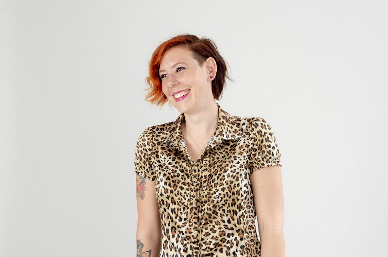 Persoon met kort, roodbruin haar glimlacht en kijkt opzij. Draagt een luipaardprint blouse en toont arm-tatoeages tegen witte achtergrond.
