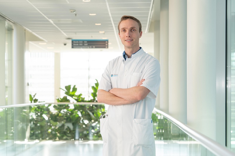 Man in witte doktersjas met gekruiste armen in lichte hal van het Catharina Hart- en Vaatcentrum, met ramen en planten.