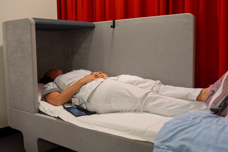 Een persoon in witte kleding ligt op een bed met handen op de buik. Naast hen liggen een tablet en koptelefoon, rode gordijn achter.