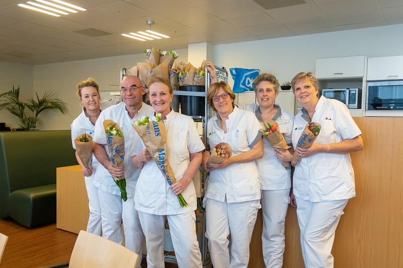 Zes mensen in witte uniformen staan samen in het Catharina Ziekenhuis, houden bloemen vast en glimlachen in een personeelsruimte.