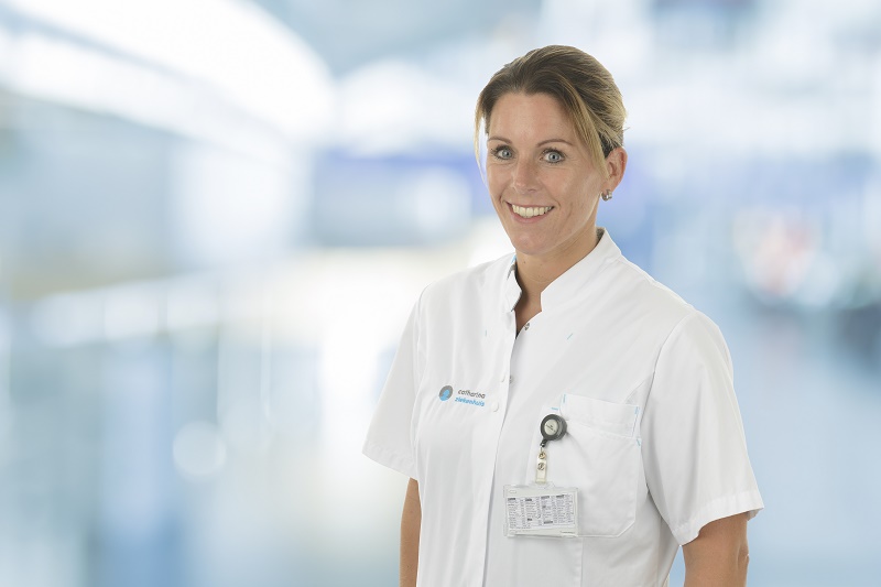 Vrouw in wit medisch uniform met naamplaatje staat glimlachend in een modern, goed verlicht zorgcentrum.