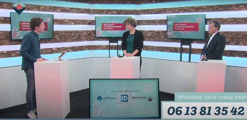 Drie mensen staan bij spreekgestoeltes in een tv-studio tijdens een coronapanel. WhatsApp-nummer voor vragen rechtsonder.