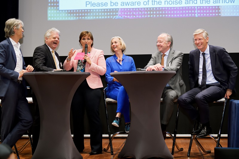 Zes zakelijk geklede mensen discussiëren rond twee statafels op een podium. Vrouw met microfoon. e/MTIC scherm op achtergrond.