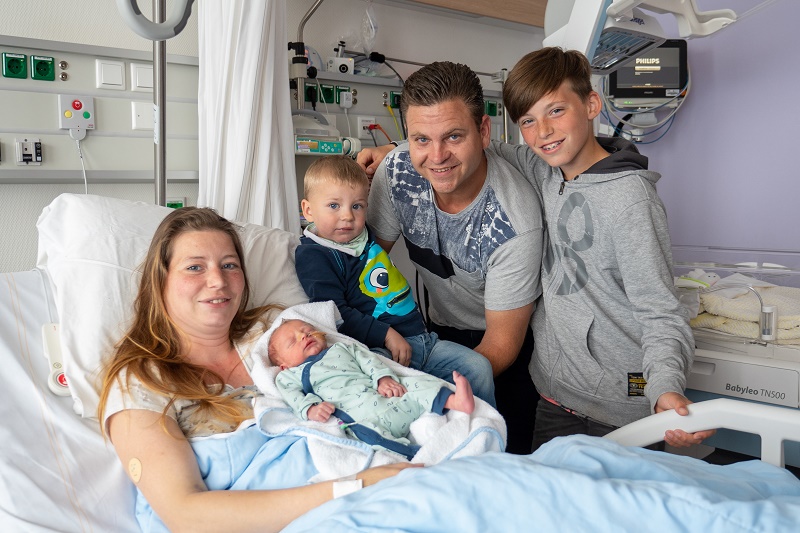 Vrouw in ziekenhuisbed met baby, jong kind naast haar. Man en oudere jongen staan erbij. Iedereen lacht naar de camera.