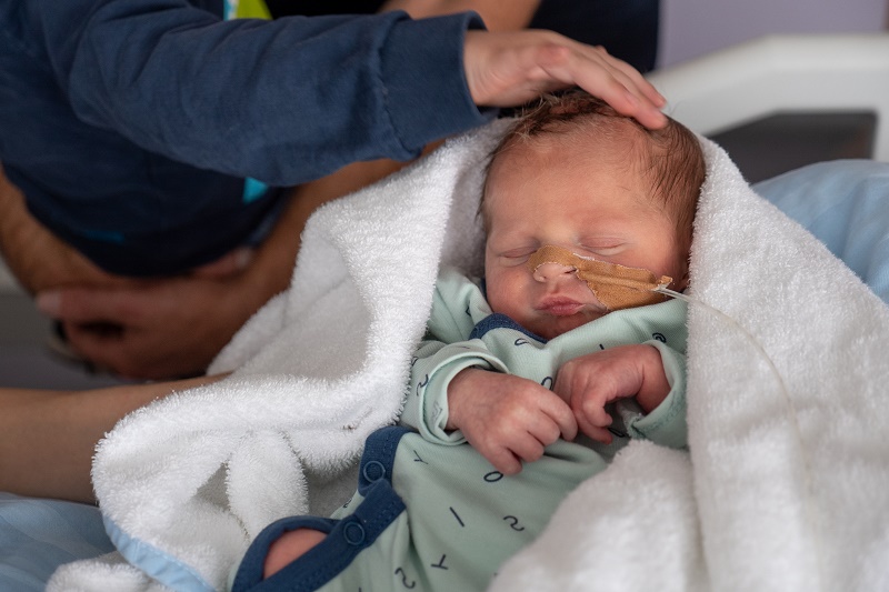 Een coronababy slaapt rustig, gewikkeld in een witte deken met een neussonde. Een hand roest op het hoofdje en een arm is zichtbaar.
