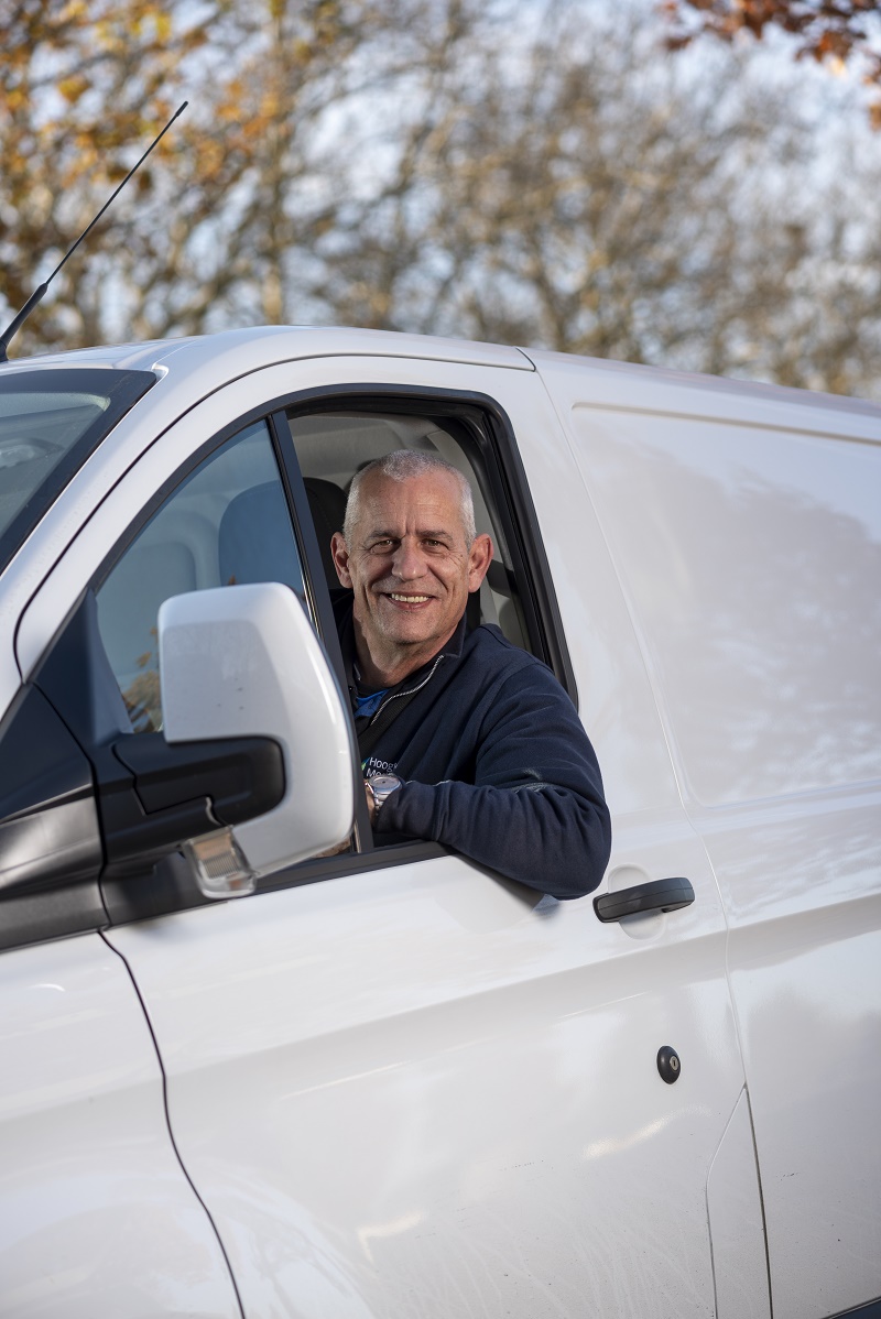 Een man met kort grijs haar zit lachend in de bestuurdersstoel van een witte bestelwagen. Er zijn herfstbomen op de achtergrond.