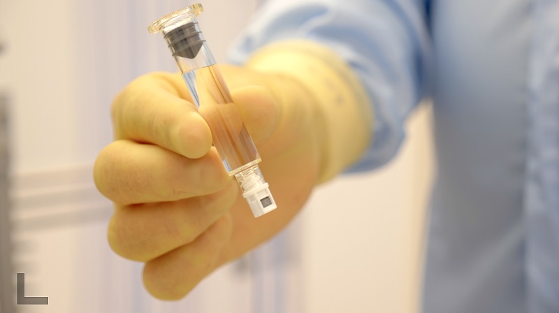 Persoon in blauwe labjas en gele handschoenen houdt glazen flesje met heldere vloeistof vast in een cleanroom van de spuitenfabriek.