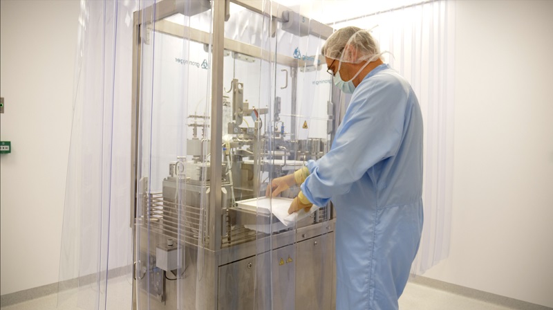 Een persoon in beschermende kleding bedient machines in een cleanroom en werkt met materialen in een transparante kast.