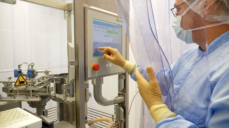 Persoon in beschermende kleding bedient touchscreen op industriële apparatuur in cleanroom van spuitenfabriek, met flacons.