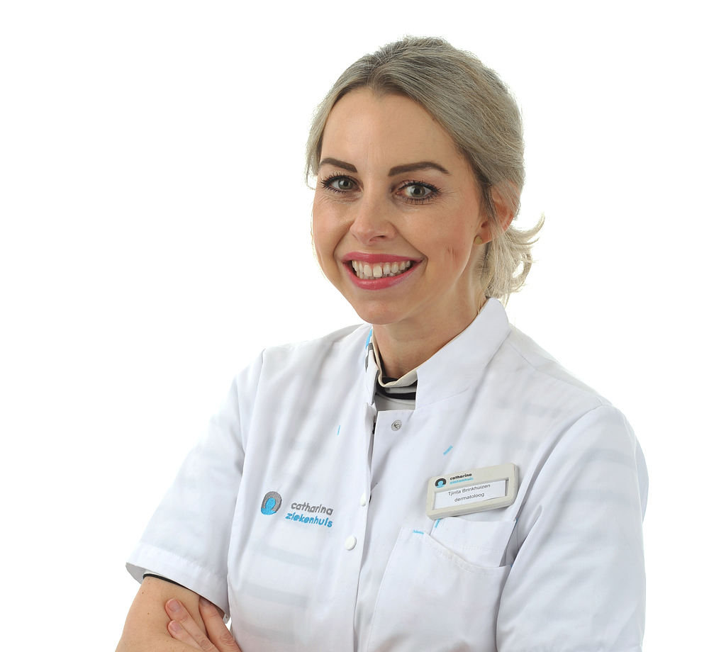 Vrouw met blond haar in wit medisch uniform en naambadge staat met gekruiste armen en glimlacht tegen witte achtergrond.