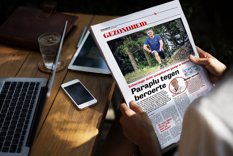 Persoon aan houten tafel met Nederlandstalige krant, dichte laptop, smartphone en glas water op tafel.