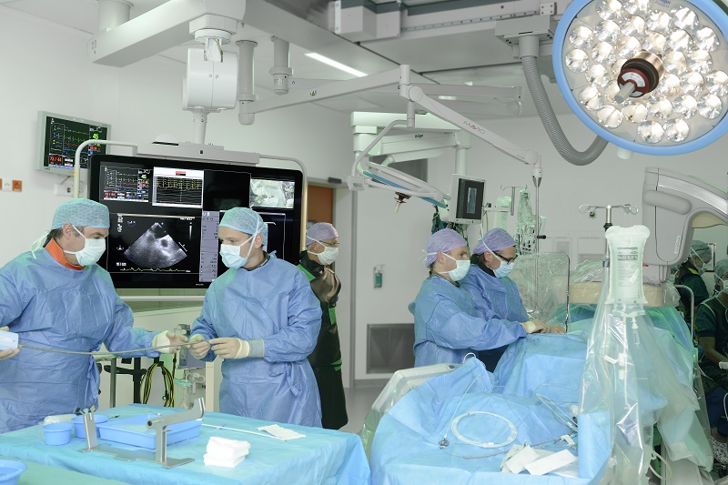 Een medisch team opereert in OK bij kransslagadervernauwing. Ze dragen schorten en maskers en letten op monitoren en instrumenten.