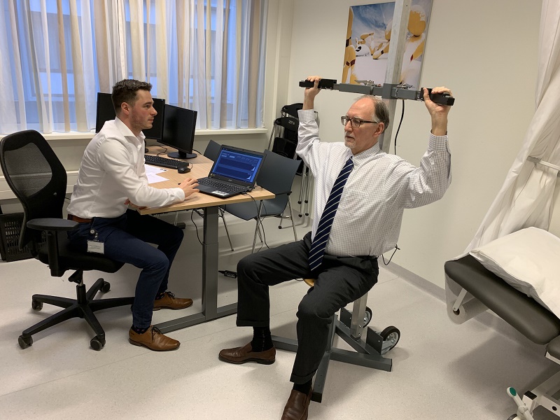 Een man in overhemd en stropdas maakt aantekeningen aan een bureau, terwijl een patiënt oefenapparatuur gebruikt in een onderzoekskamer.