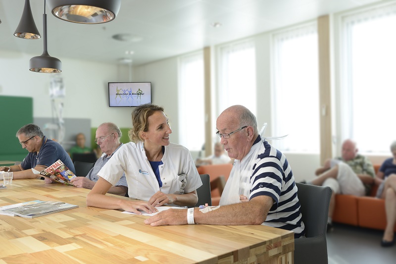 Een verpleegkundige spreekt met een oudere man aan tafel in de wachtruimte van het Catharina Ziekenhuis hart- en vaatzorg.