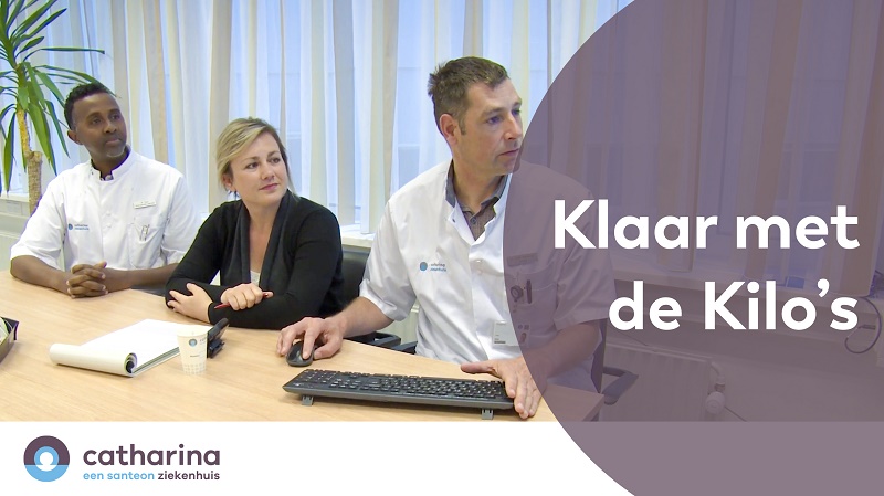 Drie personen, twee in witte uniformen en één in zwart, aan bureau met toetsenbord en papier. Tekst: "Klaar met de Kilo's".