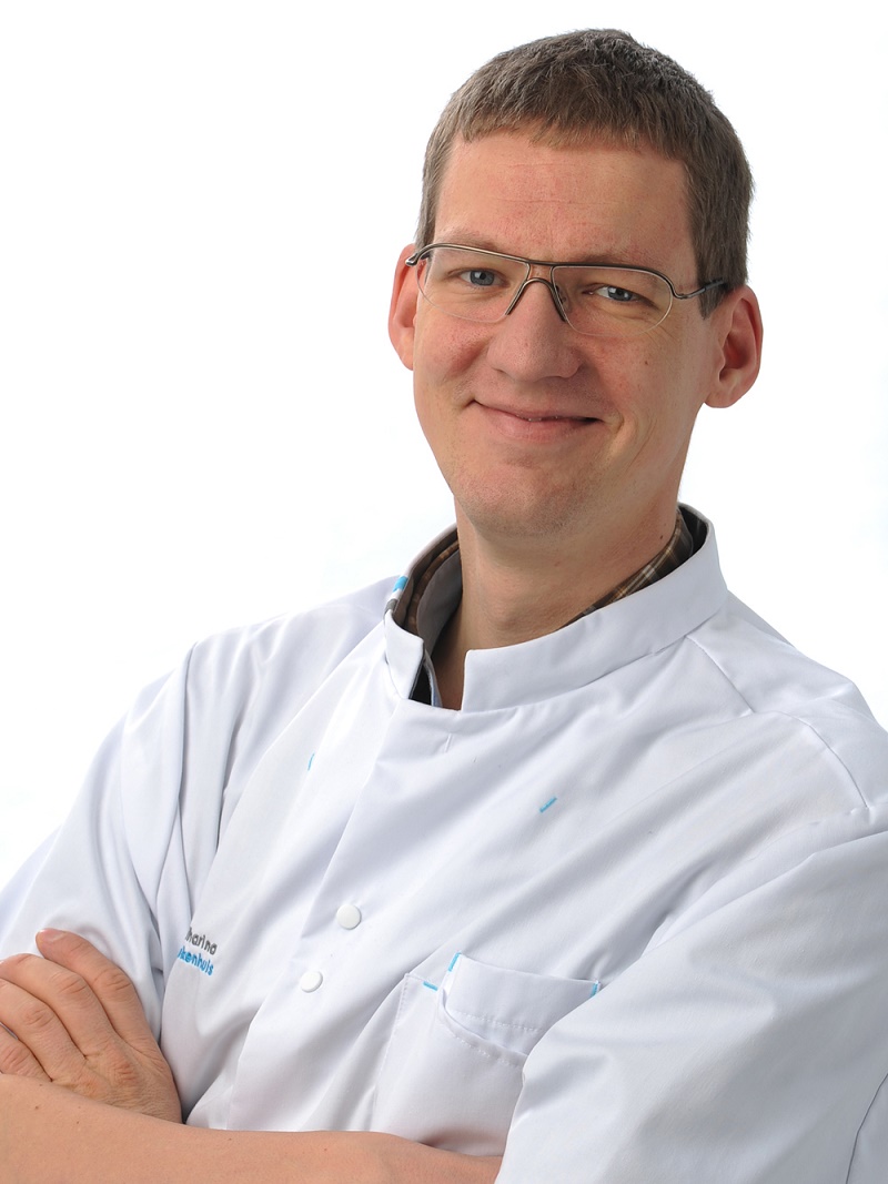 Man met bril en witte labjas staat met gekruiste armen en glimlacht naar de camera, tegen een witte achtergrond.