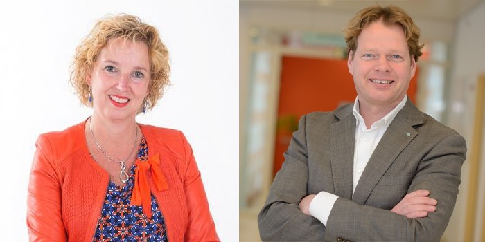 Vrouw met blond krullend haar in oranje jasje lacht links, man in grijs pak met gekruiste armen lacht rechts, binnen.