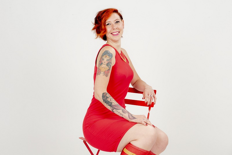 Vrouw met kort rood haar en tatoeages zit in rode jurk op een rode stoel tegen witte achtergrond en lacht naar de camera.