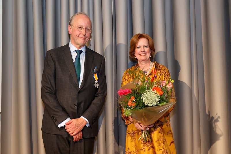 Piet Batenburg, koninklijk onderscheiden, poseert met medaille naast vrouw in gele jurk met bloemen voor grijze gordijnen.