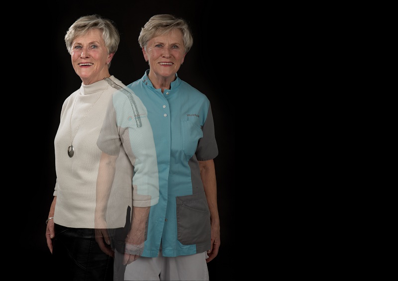 Twee oudere vrouwen met kort grijs haar staan naast elkaar, één in een witte trui, één in een blauw uniform, op zwarte achtergrond.