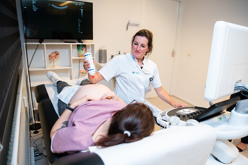 Een zwangere persoon ligt op een onderzoeksbed terwijl een zorgverlener een echo voorbereidt in een klinische ruimte.