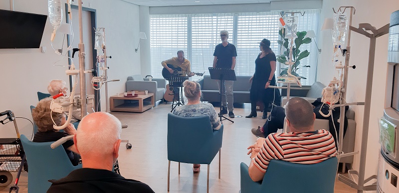 Patiënten krijgen een infuus op een moderne oncologieafdeling, terwijl drie muzikanten muziek maken tussen grote ramen.