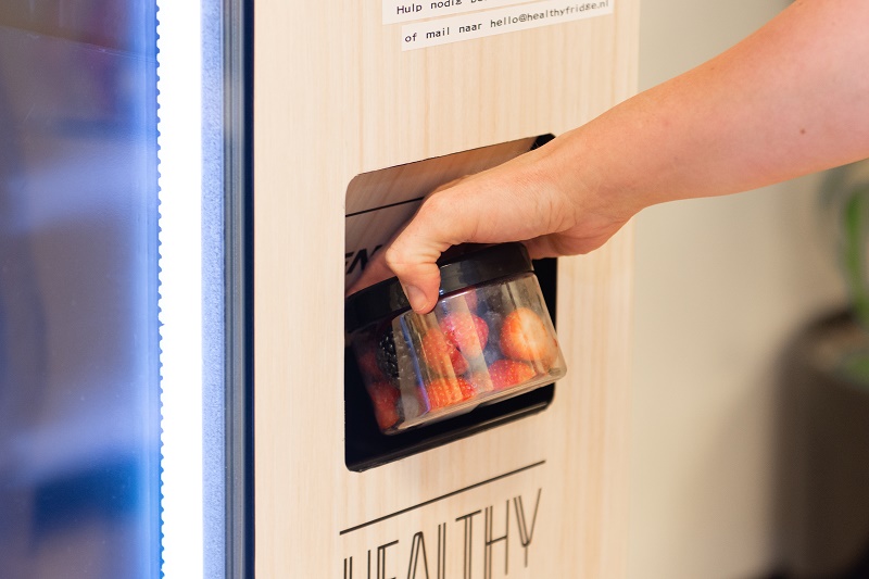 Een hand pakt een doorzichtige bak met aardbeien uit een houten snackautomaat.