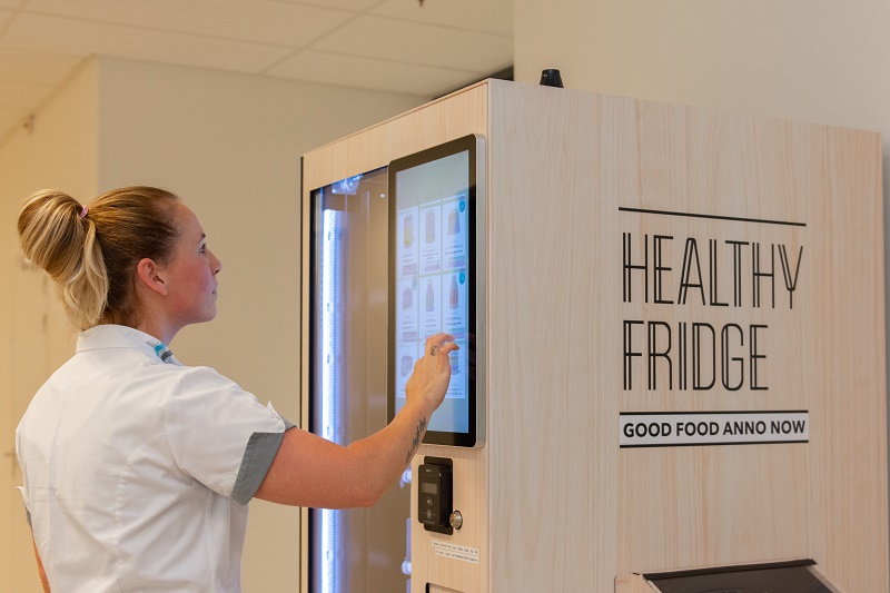 Vrouw in wit uniform bedient touchscreen van Healthy Fridge-automaat met de tekst "Good Food Anno Now" daaronder.