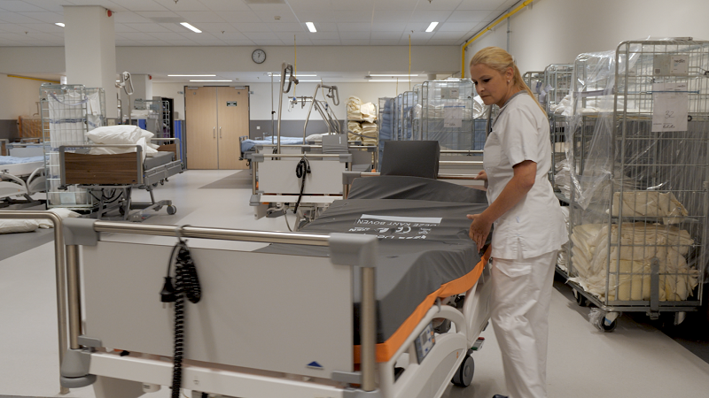 Een zorgmedewerker in witte kleding maakt een ziekenhuisbed op in een schone kamer met meerdere lege bedden en apparatuur.