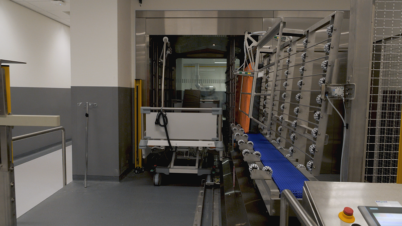 Een sterilisator met transportband en metalen rekken staat in een schone klinische ruimte met grijze vloer, muren en bediening.