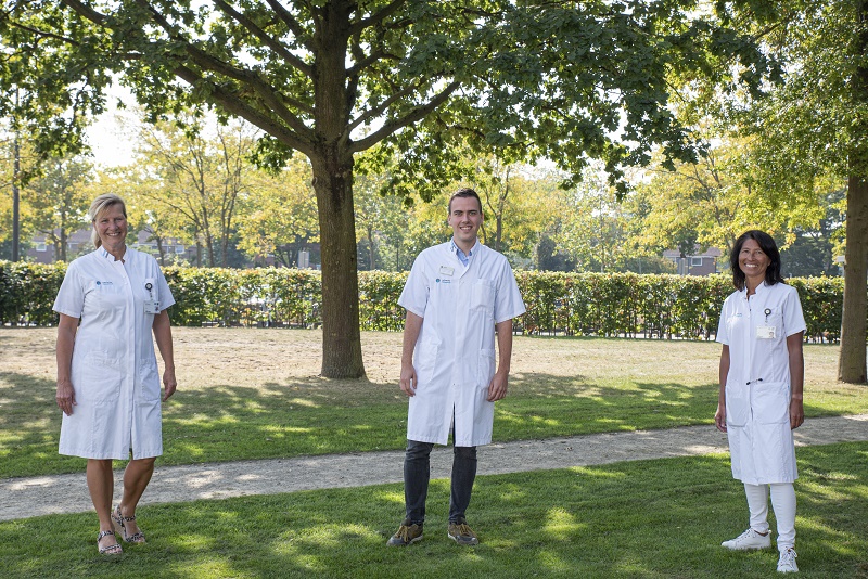 Drie mensen in witte uniformen van het Catharina Ziekenhuis staan op gras onder een boom in een park. Op de achtergrond struiken en een pad.