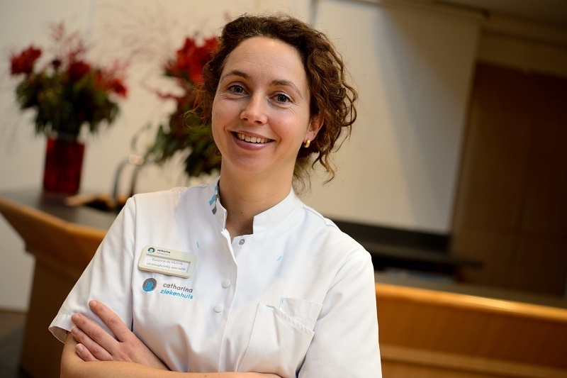 Vrouw in wit medisch uniform staat binnen bij Catharina Ziekenhuis, armen over elkaar, lachend. Bloemen en spreekgestoelte zichtbaar.