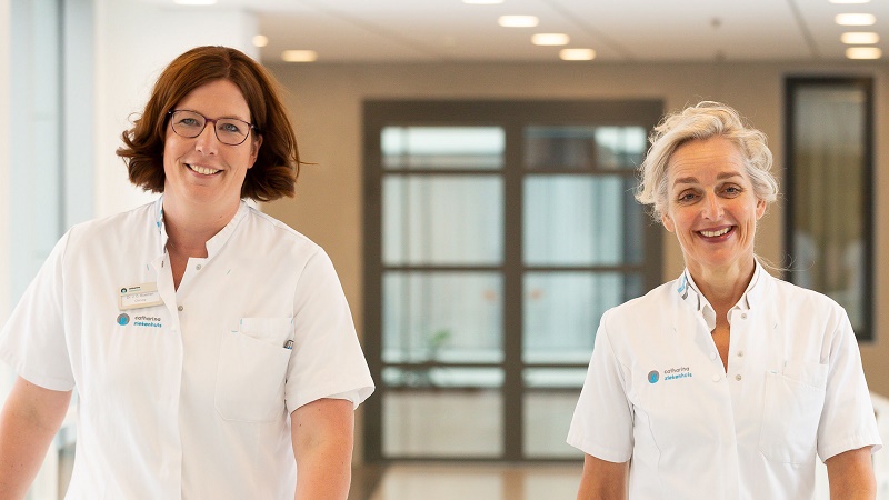 Twee vrouwen in witte medische kleding lopen glimlachend door een lichte gang, met naamkaartjes, in het natraject borstkanker.