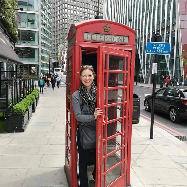 Een vrouw staat in een rode Britse telefooncel op een stoep; moderne gebouwen en verkeer op de achtergrond.