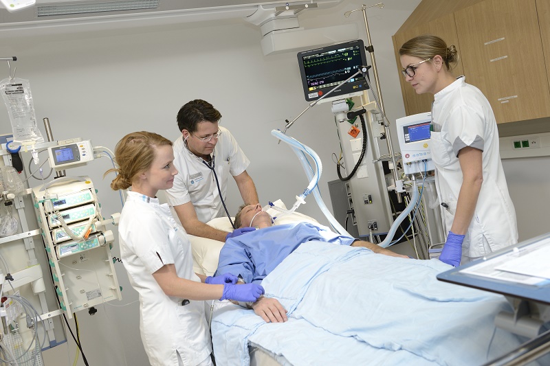 Drie zorgverleners in witte uniformen behandelen een patiënt in een ziekenhuisbed in het cathlab; medische apparatuur aanwezig.