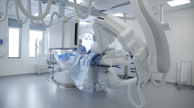 Patiënt op ziekenhuisbed in moderne cathlab met grote apparatuur; twee zorgverleners assisteren, kamer met veel licht en ramen.