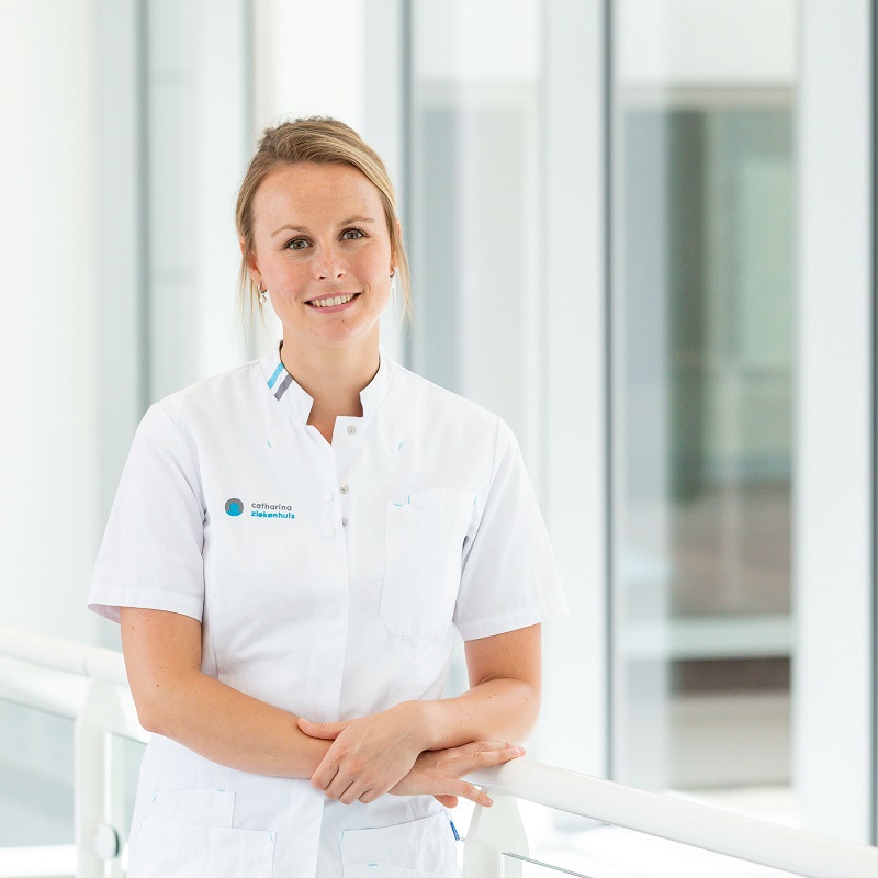 Vrouw in wit medisch uniform leunt op reling en lacht, in lichte zorgomgeving gericht op borstkanker.