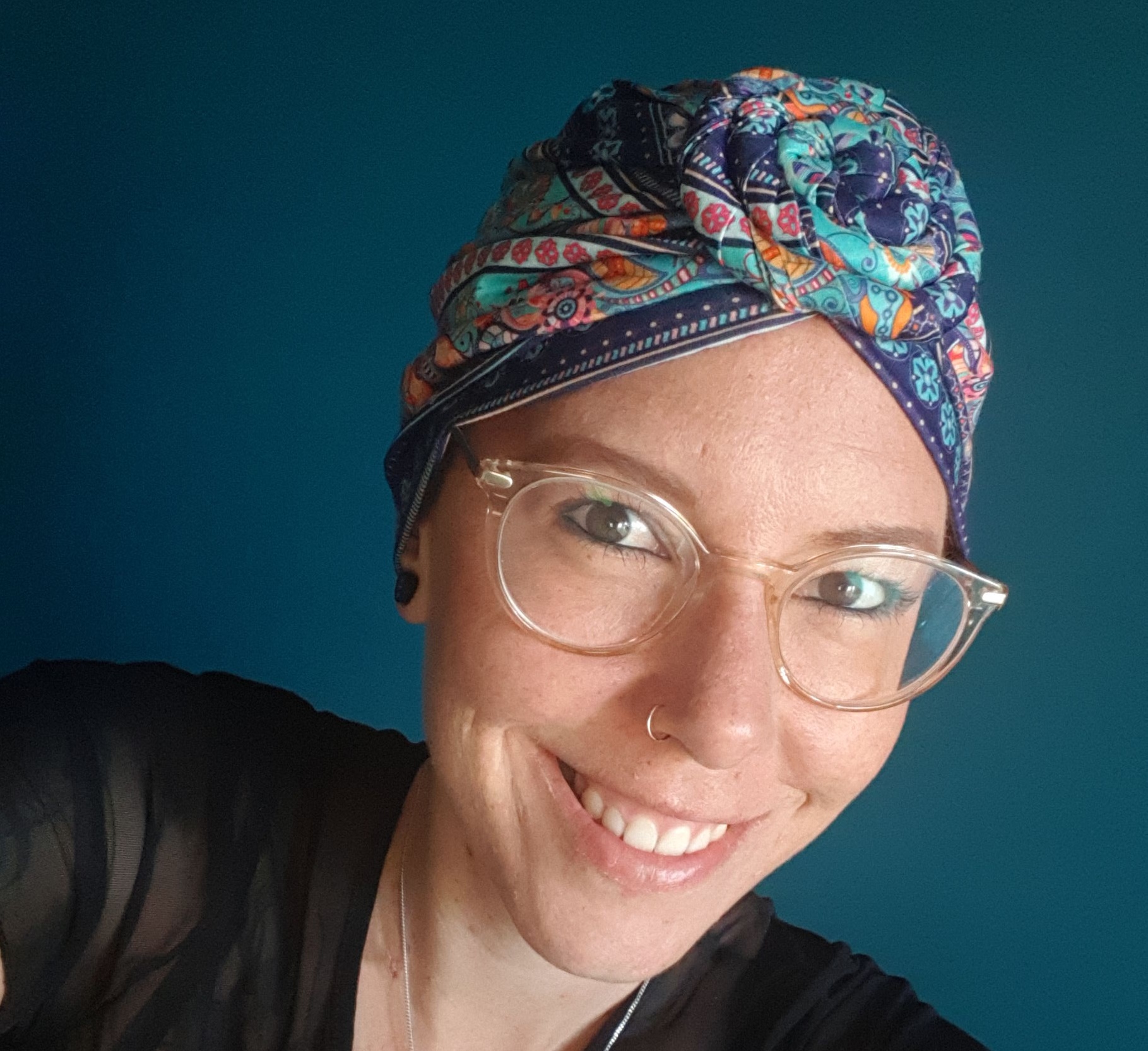 Persoon met bril en kleurrijke hoofddoek glimlacht naar de camera, tegen een effen teal achtergrond.