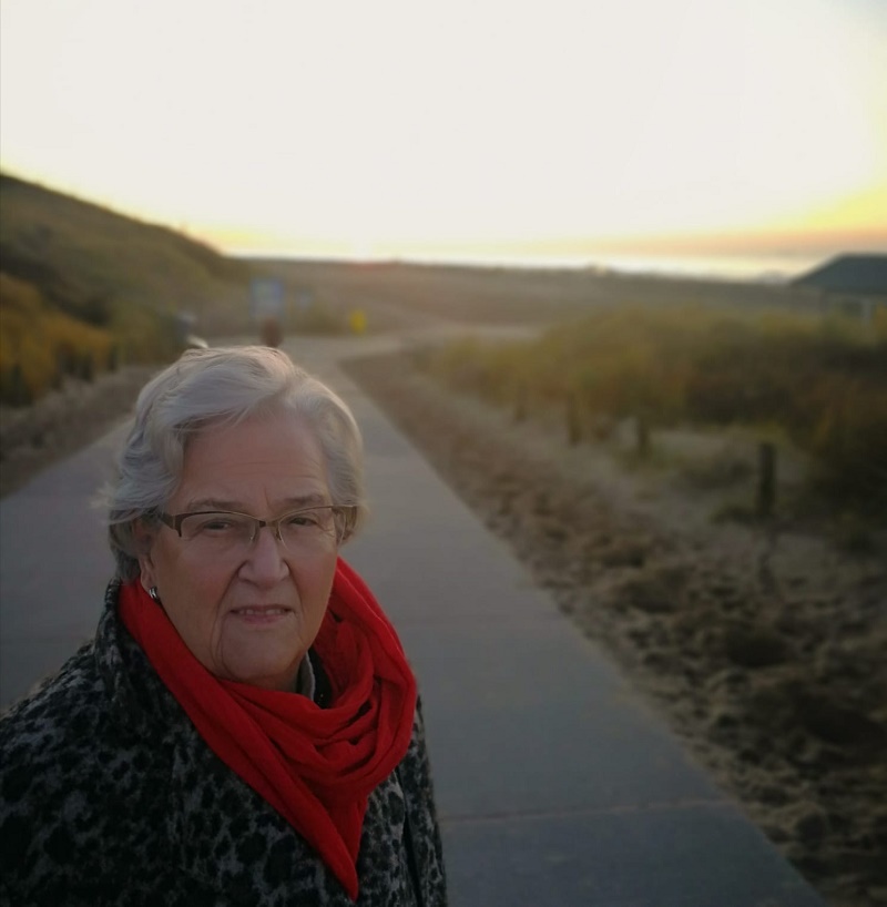 Oudere vrouw met grijs kort haar, bril en rode sjaal op een pad bij zonsondergang; zand, gras en huis op achtergrond.