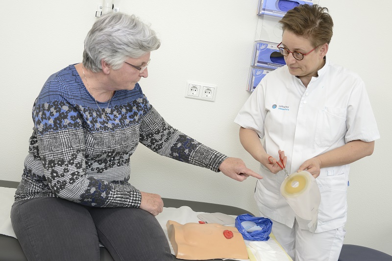Zorgmedewerker in wit uniform toont stomazakje, oudere vrouw wijst op medisch model met stomamaterialen in het Catharina Ziekenhuis.