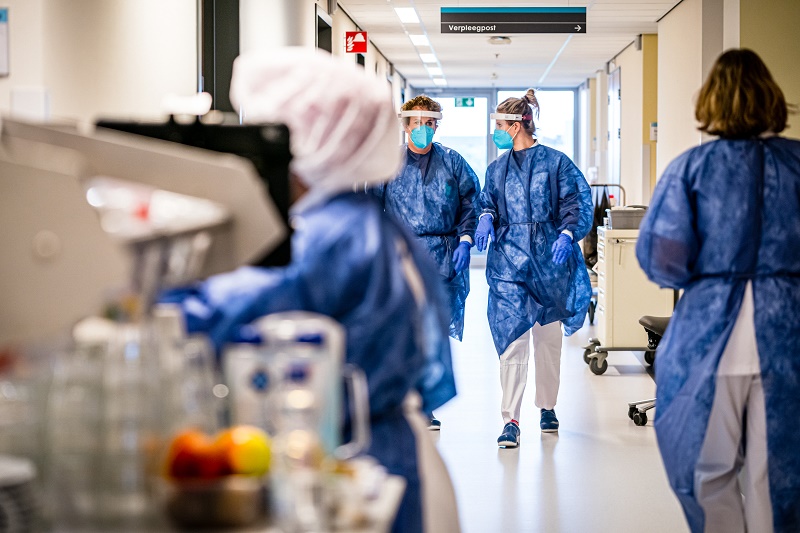 Zorgmedewerkers in blauwe beschermkleding lopen door een ziekenhuisgang tijdens de tweede golf, omringd door medische apparatuur.