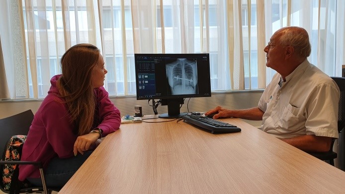 Een arts toont een borst-röntgenfoto op een computerscherm aan een Moldavisch meisje in een paarse hoodie in een verlichte kamer.