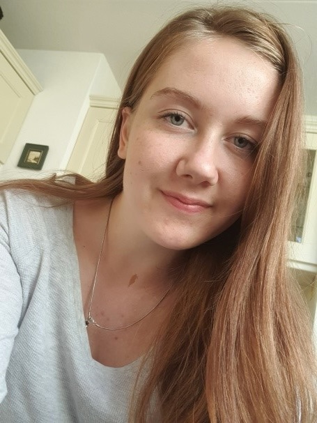 Jong Moldavisch meisje met lang lichtbruin haar en lichte huid binnen, kijkt glimlachend naar camera. Lichtgrijze top, zilveren ketting.